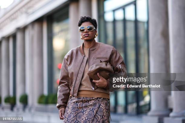 Loicka Grace wears sunglasses, golden earrings, a brown leather bomber jacket, a midi snake pattern printed leather skirt , knee high black pointed...