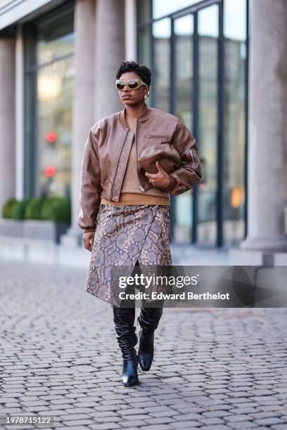 Loicka Grace wears sunglasses, golden earrings, a brown leather bomber jacket, a midi snake pattern printed leather skirt , knee high black pointed...