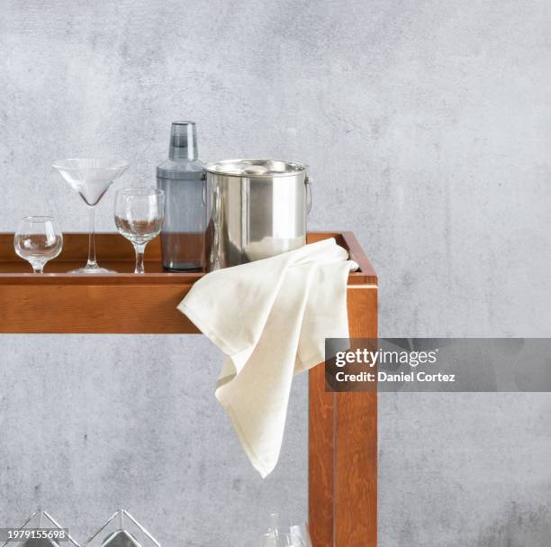 elegant home bar vignette on a walnut bar cart, stylized ice bucket, cocktail shaker, and stemware arrangement, against a textured grey wall with a linen napkin accentuating the warm wood tones. - bar cart stockfoto's en -beelden