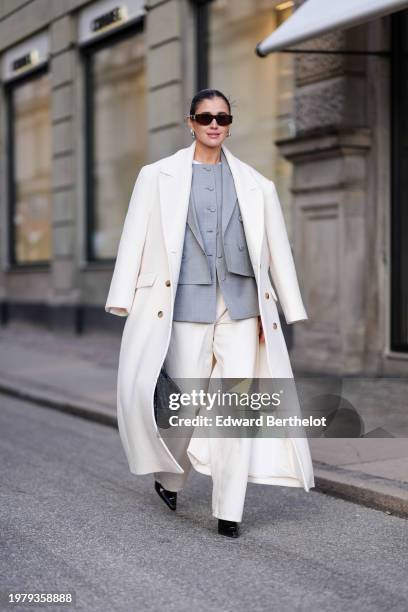 Darja Barannik wears sunglasses, golden earrings, a white oversized long coat, a gray oversized blazer jacket , white flared suit pants , pointed...