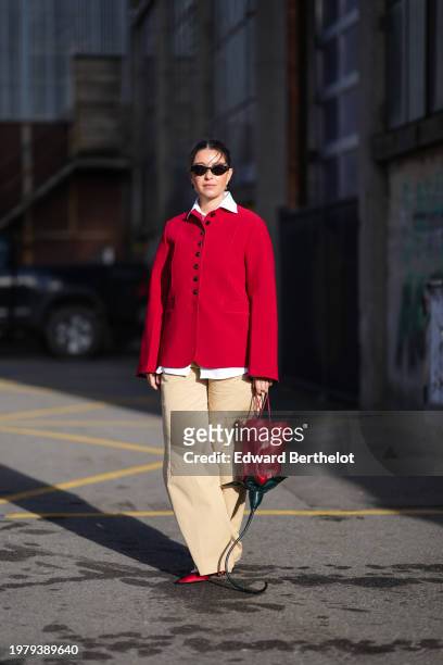 Guest wears sunglasses, a shirt, a red oversized blazer jacket , beige flared pants a red leather bag shaped as a rose / flower, red shoes outside...