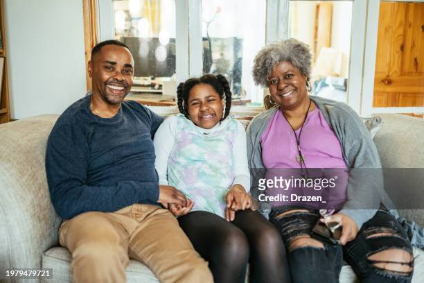 mature african-american parents sitting on couch with teen daughter. family looking at camera and smiling - afro caribbean portrait stock pictures, royalty-free photos & images