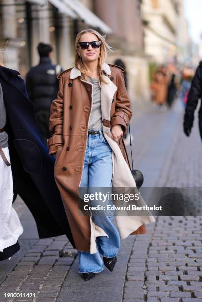 Guest wears sunglasses, a brown leather long trench coat, a gray top, thin belt, blue denim jeans pants , pointed shoes, outside Mark Kenly Domino...