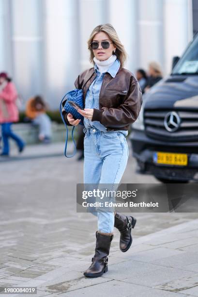 Guest wears sunglasses, a white turtleneck pullover, a blue denim shirt, a blue woven leather bag, blue denim jeans pants , brown leather boots...