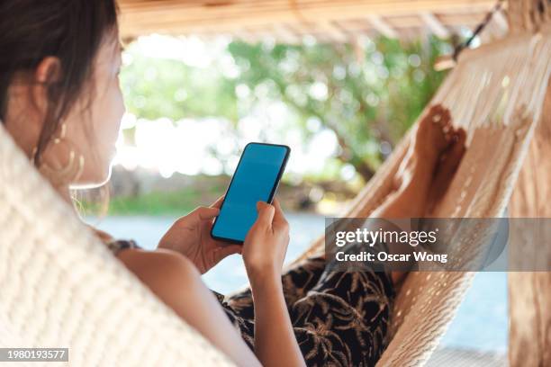 over the shoulder view of young asian woman using smartphone while relaxing on hammock in tropical resort - over the shoulder view stock pictures, royalty-free photos & images