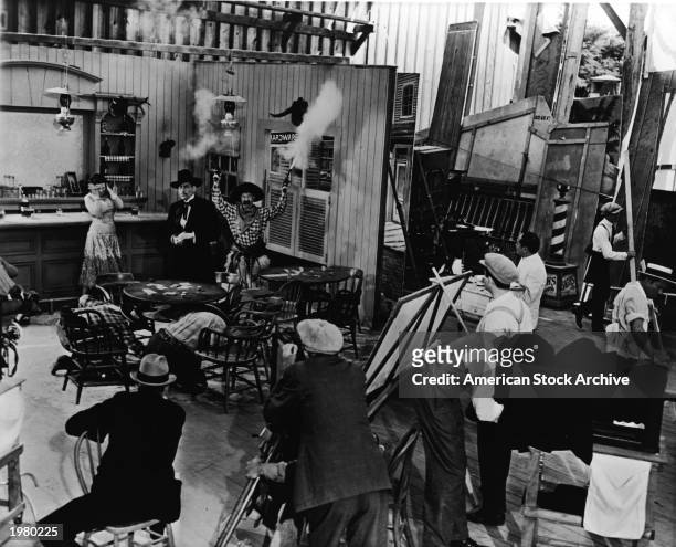 An actor wearing a cowboy costume shoots two pistols in the air as other actors react on an Old West saloon studio set during the production of a...