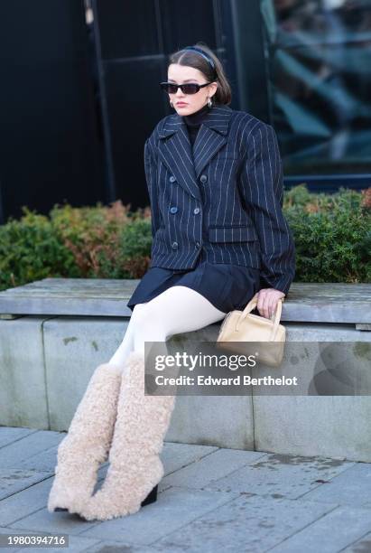 Guest wears a headband, sunglasses , earrings, a black turtleneck pullover, a black and white striped oversized blazer jacket, a beige leather bag, a...