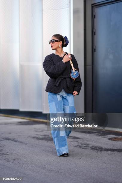 Guest wears sunglasses, golden earrings, a necklace , a scrunchie, a blue monogram logo printed Vuitton bag, a black oversized padded jacket , blue...