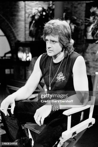 American Rock and Pop musician Eddie Money holds a cigarette as he sits on director's chair during an MTV interview at Teletronic Studios, New York,...