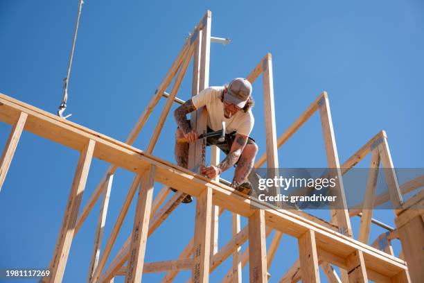 carpinteiro sentado em cima de parede áspera emoldurada pregando em uma treliça de telhado sob um céu azul - armação de construção - fotografias e filmes do acervo