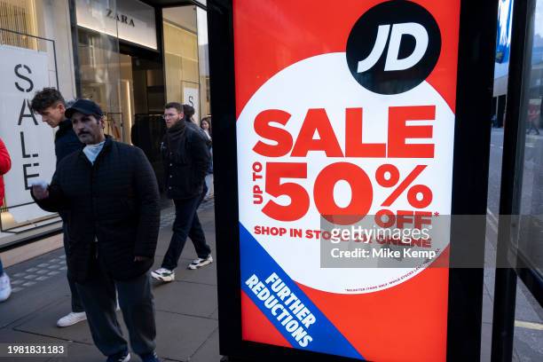 People out shopping on Oxford Street walk past large scale January sale signs in red and white for major high street clothing retail shops including...