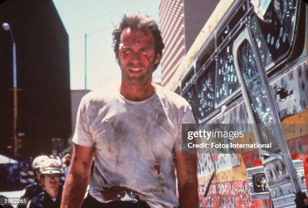 American actor and director Clint Eastwood walks on a city street beside a bullet-riddled bus, with dirt and blood staining his face and shirt and a...