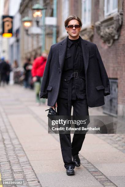 Guest wears sunglasses, a dark black jacket , a black shirt, suit pants, black leather loafers shoes, a mini Hermes bag, outside The Garment, during...