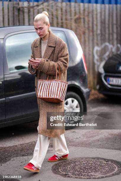 Guest wears earrings, a brown long coat, a basket bag, beige flared pants , red Adidas sneakers shoes , holds a mobile phone, outside Skall Studio,...