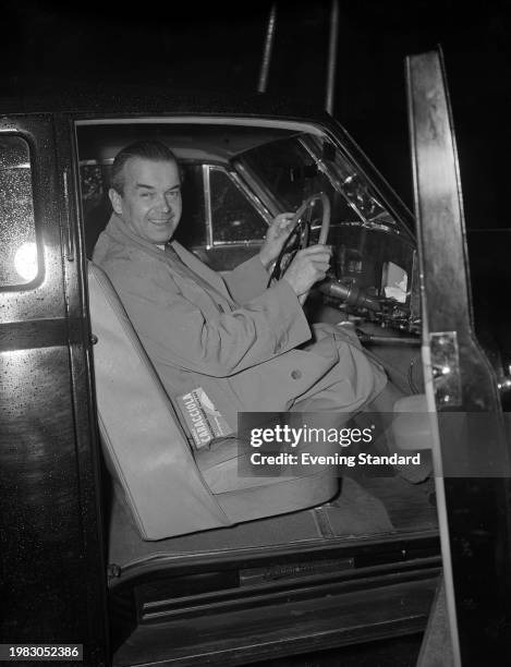 German racing driver Rudolf Caracciola smiling as he sits at the wheel of a car, a copy of his autobiography sticking out from the pocket of his...