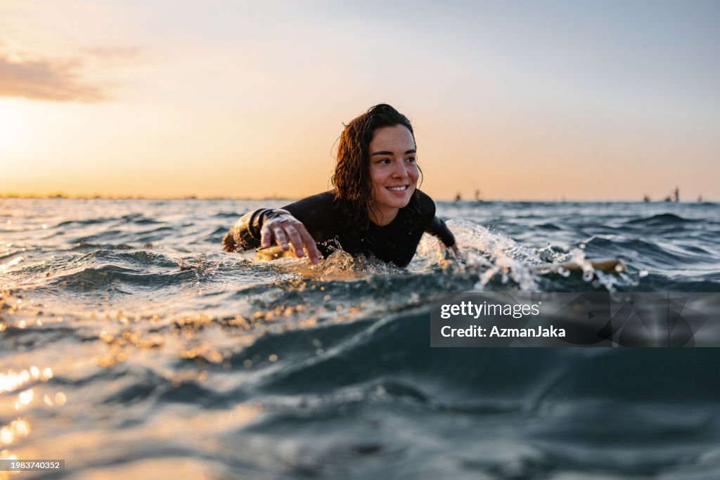 Attraktive junge erwachsene Surferin auf einem Surfbrett, die versucht, eine Welle während des Sonnenuntergangs zu fangen