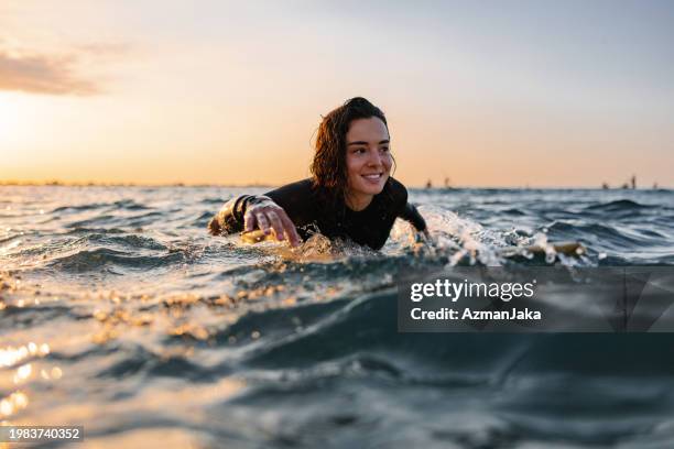 attraktive junge erwachsene surferin auf einem surfbrett, die versucht, eine welle während des sonnenuntergangs zu fangen - adrenalin stock-fotos und bilder