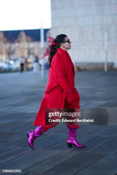 Guest wears a red bow hair attachment detail, sunglasses, earrings, a red long coat , purple shiny pointed boots , outside OperaSport, during the...