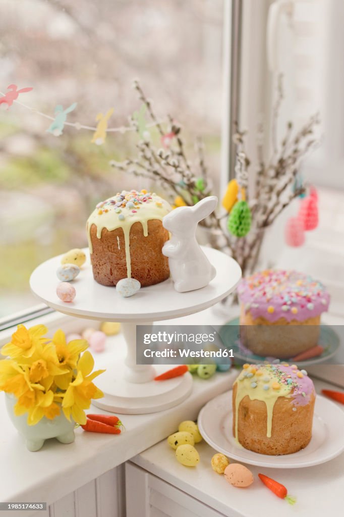 Cakes decorated with flowers and eggs near Easter bunny toy on window sill