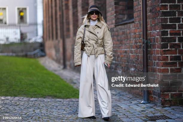 Guest wears a hat, a white turtleneck pullover , a beige short trench coat, white palazzo flared pants, a brown bag outside Herskind, during the...