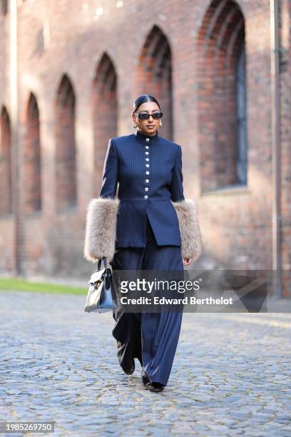 Guest wears golden earrings, sunglasses , a navy blue military jacket with golden buttons and beige fluffy sleeves, matching flared pants, outside...