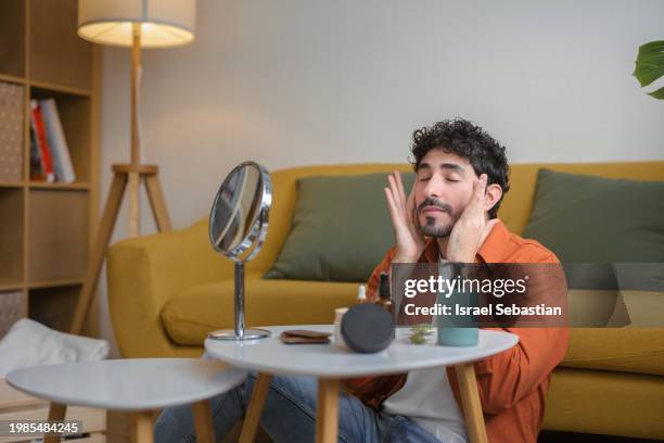 man sitting on sofa applying facial cream on his cheek. - facial cleanser stock pictures, royalty-free photos & images