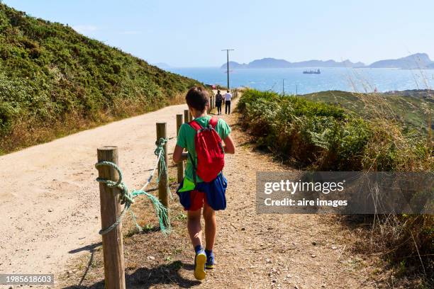 a boy walking backwards along a dirt road with a protective fence carrying a red backpack in the background part of the cies islands can be seen cape home, spain. - vigo stock pictures, royalty-free photos & images