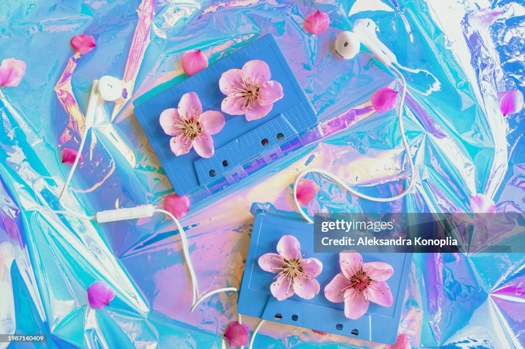 Two analog audio cassette with wired headphones and pink peach tree flowers, spring bloom on crumpled holographic foil background pattern texture. Flat lay, top view, copy space.