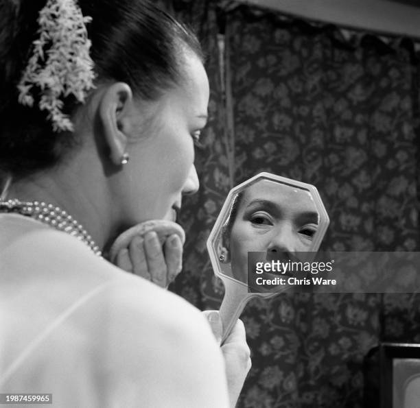 Burmese-born Millicent Spencer looking in a handheld mirror while applying a homemade sandalwood paste to her face, Stockell, London, November 1955.