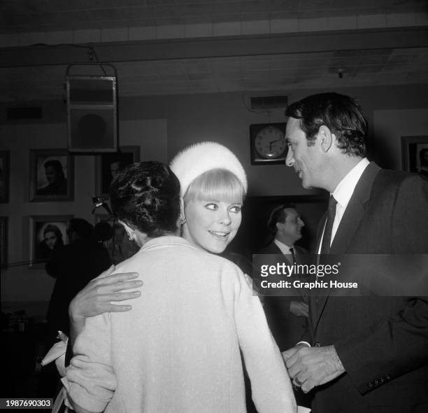 German actress Elke Sommer, wearing a white fur hat, smiles as she embraces a person beside her husband, American columnist and author Joe Hyams, who...