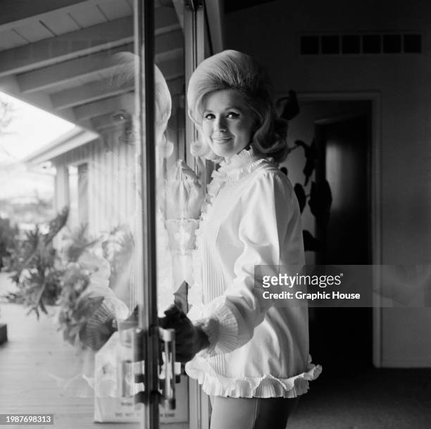 American actress and singer Connie Stevens, wearing a white top, trimmed with frills on the collar, cuffs and hem, poses against patio doors, at home...