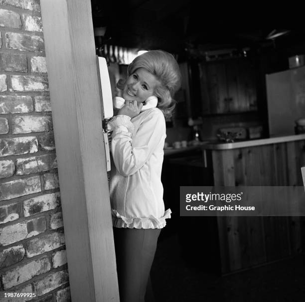 American actress and singer Connie Stevens, wearing a white top, trimmed with frills on the collar, cuffs and hem, poses using a telephone, at home...