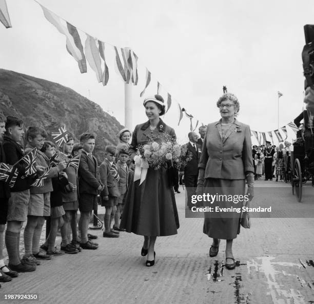 Queen Elizabeth II carries a bouquet of flowers while walking with Sybil Hathaway, Dame of Sark in front of a line of boys waving Union Jack flags...