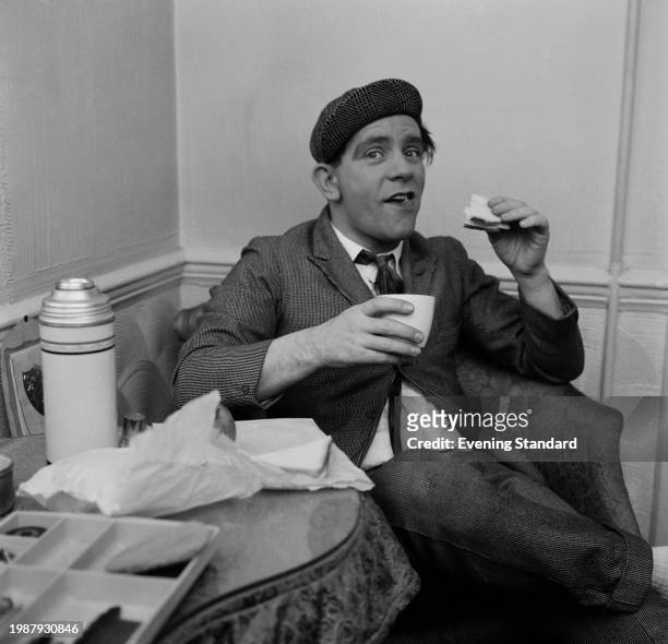 British comedian and actor Norman Wisdom eating a sandwich and holding a drink backstage, January 25th 1956.