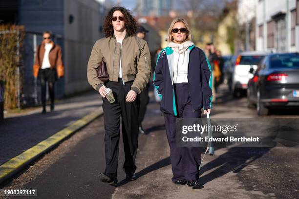 Pim Spoor wears sunglasses, a khaki bomber jacket , a brown leather bag, black pants ; guest wears sunglasses, a white wool balaclava, a navy blue...