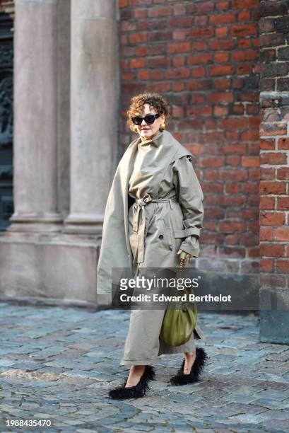Renia Jaz wears sunglasses, golden earrings , a beige oversized trench long coat with integrated scarf, a green khaki leather bag, fluffy shoes, ,...