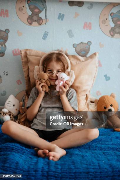 cute girl sitting on bed at home with cuddly toys and ear muffs - animal representation stock pictures, royalty-free photos & images