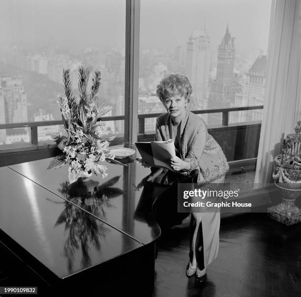 American comedian and actress Lucille Ball, leaning against a grand piano, a script in her hand, before a window in her New York Hilton apartment,...