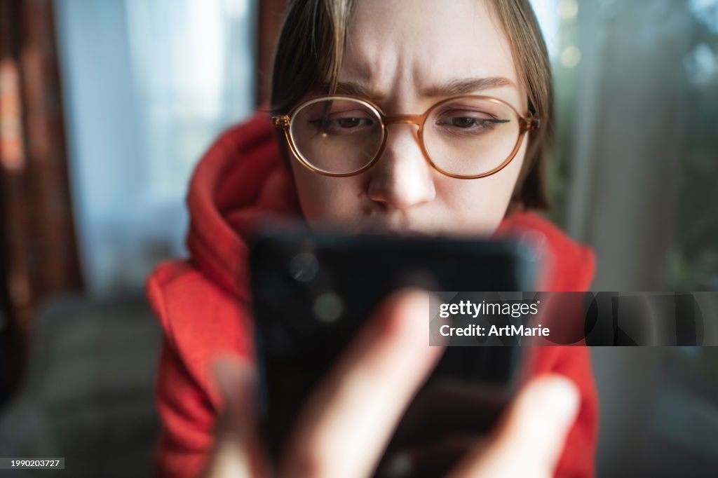 Concerned, sad, tired and worried young woman with smartphone at home reading some bad news or having problems or fraud