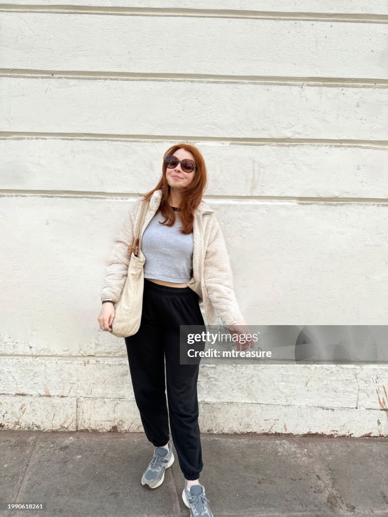 Image of attractive, redheaded woman wearing sunglasses, fluffy jacket, jogging trousers, crop top, tote bag on shoulder, smiling looking at camera, white wall background, focus on foreground, copy space