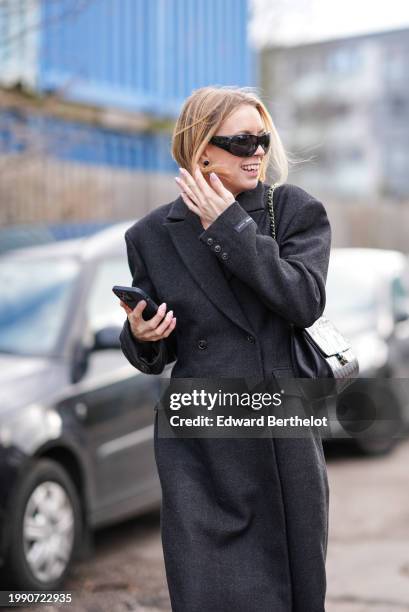 Guest wears earrings, sunglasses, a black double breasted long winter coat , a black leather Chanel bag, holds a mobile phone, outside Skall Studio,...