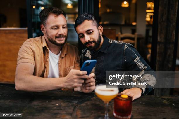 smiling gay couple using a phone over drinks on a bar patio - day in the life series stock pictures, royalty-free photos & images