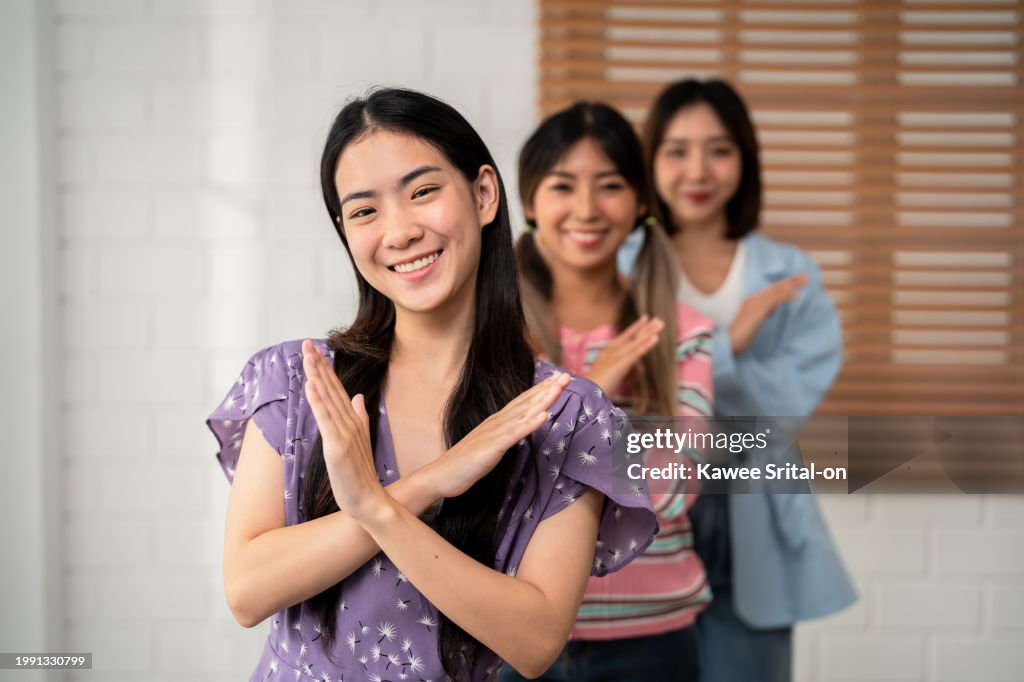 Portrait group of women friends making stop sign with crossed hands. Attractive young girls making x sign or gesturing stop for denying, refusing any action then smiling, looking at camera in house.