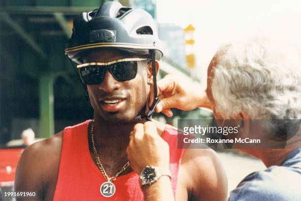 Deion Sanders has a bicycle helmet adjusted while wearing a red tank top, a necklace with a 21 medallion and sunglasses on the set of a Nike "Deion...