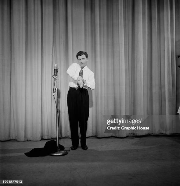 American comic actor Eddie Bracken, his tongue lolling as he grips his right wrist with his left hand in a shirt and tie, his jacket on the floor by...