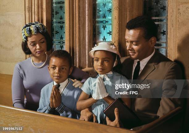 Mother and father sit on either side of their son and daughter, who both hold their hands together in prayer, as they attend church, United States,...
