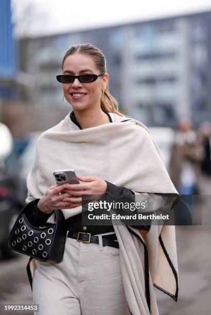 Guest wears sunglasses, a long white scarf / poncho , a black leather bag, a leather belt, white suit pants, outside Skall Studio, during the...