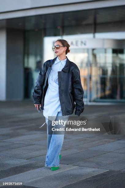 Guest wears sunglasses, earrings, a pastel pale blue long shirt, a black oversized leather jacket , blue denim jeans pants outside OperaSport, during...