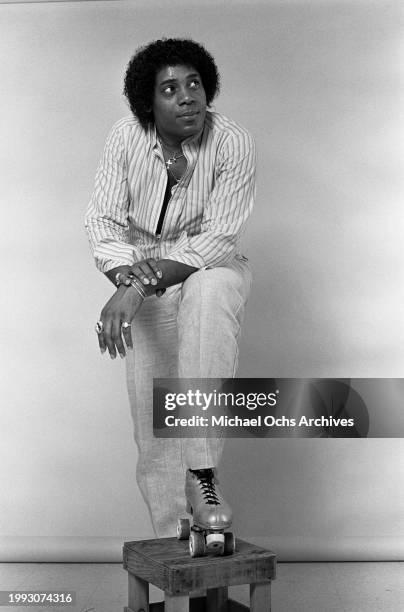 American singer Sterling Harrison wearing a striped shirt, light trousers, and roller skates, poses with his left foot on a stool in a studio...