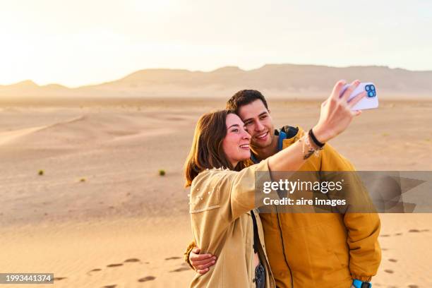 portrait of a young couple taking a selfie with their phone in the zagora desert, front view - geology technology stock pictures, royalty-free photos & images